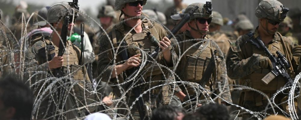 TOPSHOT - US soldiers stand guard behind barbed wire as Afghans sit on a roadside near the military part of the airport in Kabul on August 20, 2021, hoping to flee from the country after the Taliban's military takeover of Afghanistan. (Photo by Wakil KOHSAR / AFP) (Photo by WAKIL KOHSAR/AFP via Getty Images)